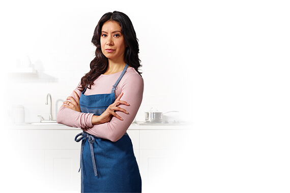 A mom wearing an apron standing in front of a kitchen counter with her arms crossed.